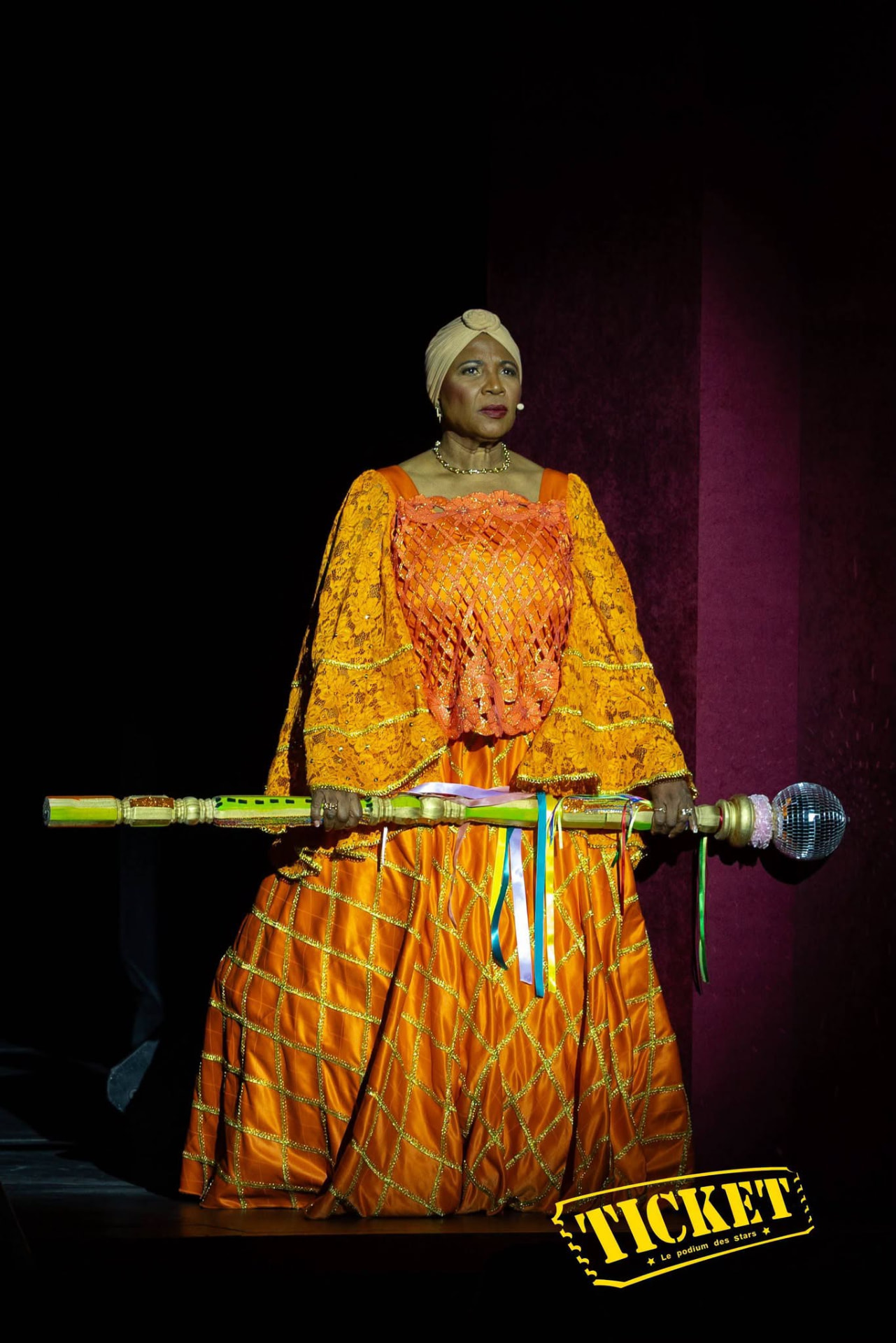 A performer in an orange gown holding a ribboned ceremonial staff