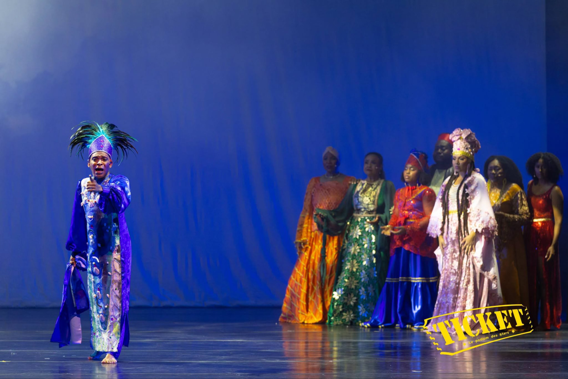 A performer in a plumed blue headdress before the ensemble in colorful gowns